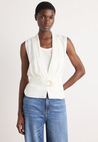 White sleeveless vest with a draped neckline, cinched waist featuring a gold buckle. Paired with high-waisted blue denim jeans.