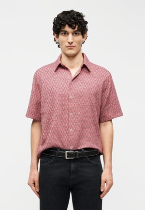 Mand med kort krøllet hår iført en lyserød mønstret kortærmet skjorte med knapper, stoppet ned i sorte jeans med sort bælte.