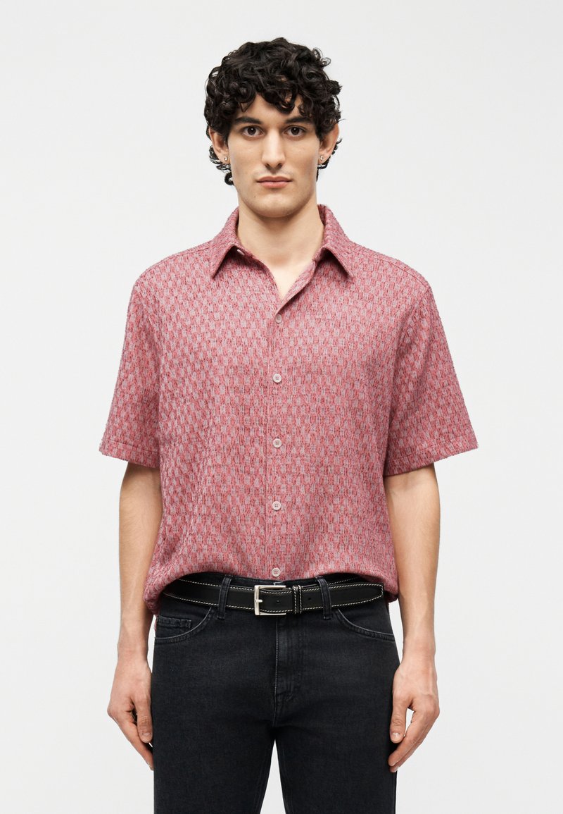 Άνδρας με κοντά σγουρά μαλλιά που φοράει ροζ πουκάμισο με κοντά μανίκια και σχέδια, κρυμμένο μέσα σε μαύρο τζιν με μαύρη ζώνη.