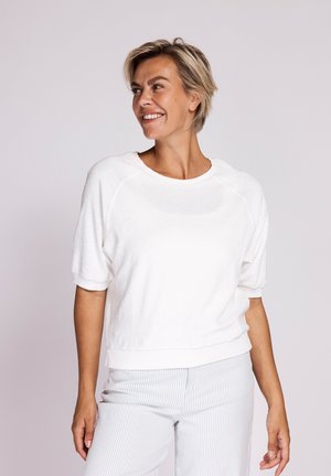 Lächelnde Frau mit kurzen blonden Haaren, die ein weißes kurzärmeliges Oberteil und hellgraue gestreifte Hosen trägt, blickt nach links.