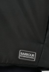 Ύφασμα μαύρου μπουφάν με λεπτή υφή, με ένα ορθογώνιο ετικέτα «Barbour International» σε λευκό και γκρι ραμμένη στο πλάι.