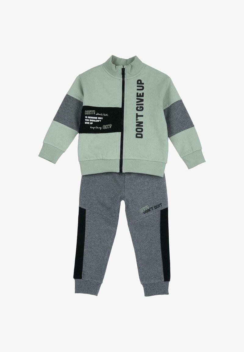 Dvoudílný teplákový komplet s mátově zelenou bundou se černými a šedými akcenty a šedými kalhotami. Bunda má textové vzory, vysoký límec a zip.