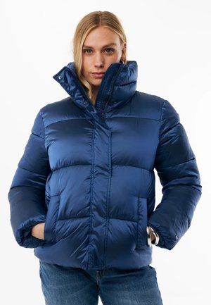 Frau mit blondem Haar, die eine blaue Steppjacke mit hohem Kragen trägt und die Hände in den Taschen hat, kombiniert mit blauen Jeans vor weißem Hintergrund.