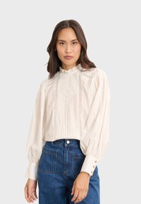 Hellbeige Bluse mit hohem Rüschenausschnitt, aufwendiger Stickerei und Falten-Design; kombiniert mit hochgeschnittenen blauen Jeans.