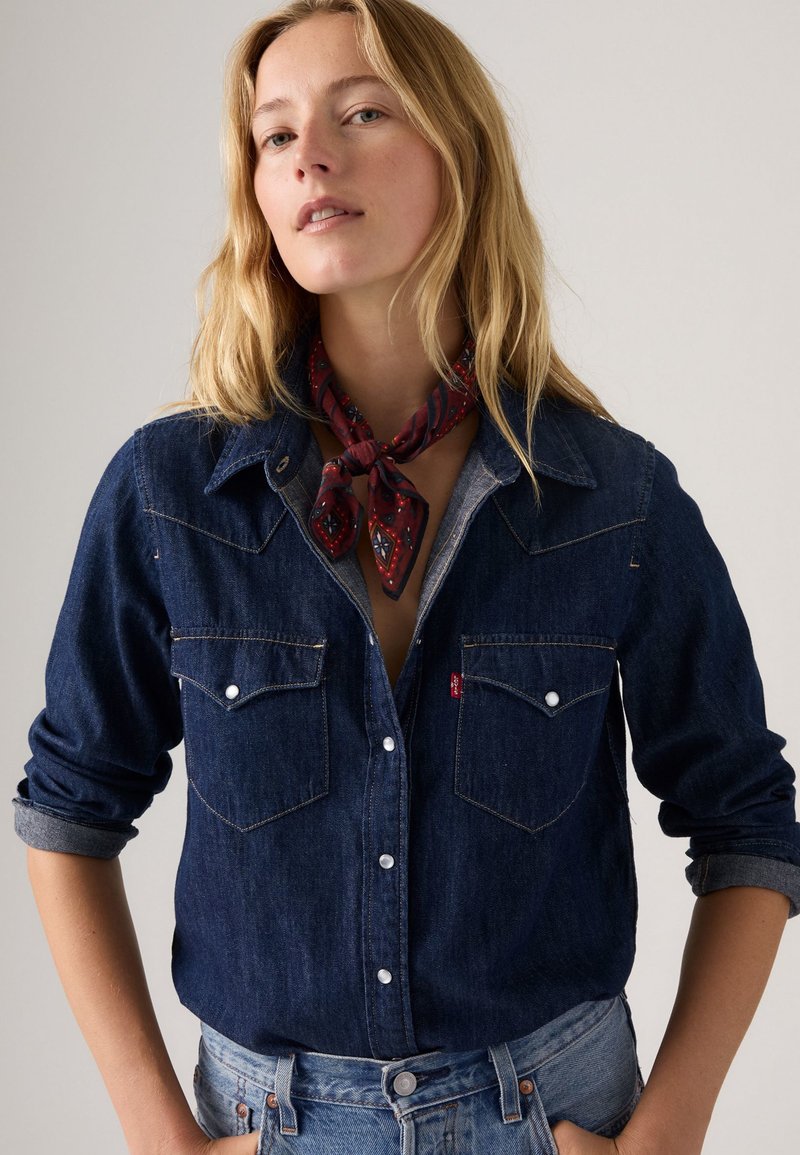 Camicia in denim scuro con bottoni a perla, due tasche sul petto e maniche arrotolate. Sciarpa rossa e a fantasia annodata attorno al collo per un tocco di stile.