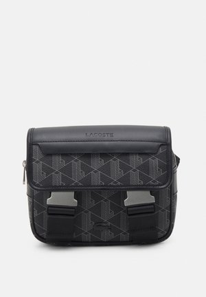 Černá crossbody taška Lacoste s geometrickým vzorem, klopou s raženým logem, dvěma kovovými přezkami a nastavitelným ramenním popruhem.