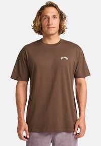 Brun bomulds t-shirt med rund hals og korte �ærmer, med et hvidt logo på brystet. Lige snit.