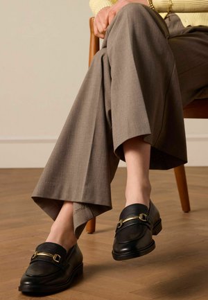 Schwarze Lederslipper mit goldfarbenen Beschlägen, getragen zu grauen, weit geschnittenen Hosen, die eine glatte Textur und ein minimalistisches Design zeigen.