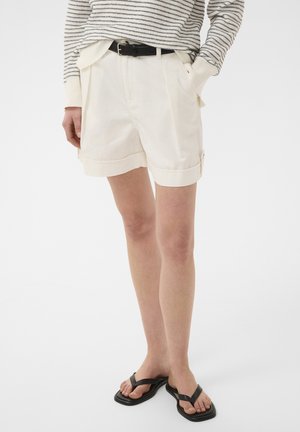 Persoon draagt witte geplooide shorts met omgeslagen zomen, een zwarte riem, een gestreepte top met lange mouwen en zwarte sandalen met vierkante neus.