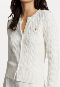 Mujer vestida con un cárdigan blanco de punto trenzado con botones y un pequeño logo bordado en el pecho, combinado con pantalones blancos.