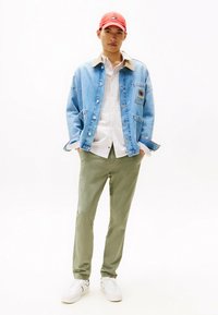 Giacca di jeans azzurro chiaro con colletto marrone, camicia bianca, pantaloni verde oliva e sneakers bianche. Cappellino rosso brillante. Look casual e sovrapposto.