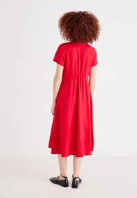 Κόκκινο, κοντομάνικο φόρεμα με ελαστική μέση, που φτάνει μέχρι το μέσο της γάμπας. Το ύφασμα φαίνεται λείο. Συνδυάζεται με μαύρες, επίπεδες γόβες.