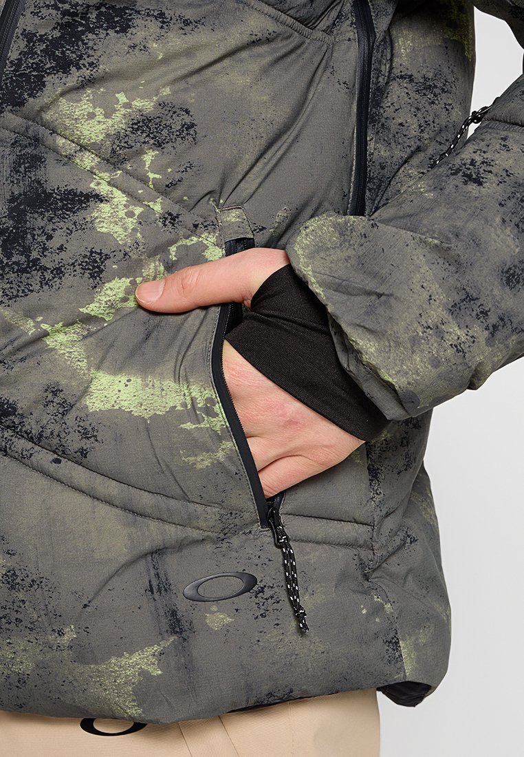 Veste rembourrée de couleur gris foncé avec des motifs de éclaboussures vertes. Elle dispose d'une poche zippée, de poignets élastiques et d'un cordon de serrage à l'ourlet.