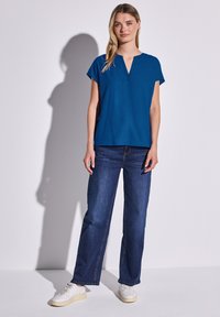 Top blu corto a maniche corte con scollatura a V, abbinato a jeans in denim scuro a gamba dritta e sneakers bianche su uno sfondo chiaro.