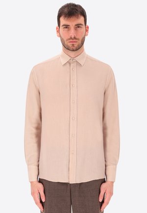 Uomo con capelli corti e scuri che indossa una camicia beige a maniche lunghe con bottoni e pantaloni marroni, in piedi davanti a uno sfondo chiaro e uniforme.