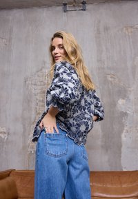Marineblaue Bluse mit Blumenmuster und weißer Stickerei, mit gewellten Kanten, kombiniert mit hellblauen Jeans mit weitem Bein. Im Hintergrund ist eine strukturierte Wand zu sehen.