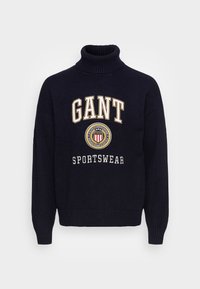 Maglione a collo alto lavorato a maglia blu navy con il testo bianco "GANT" e "SPORTSWEAR", un design del logo circolare al centro e polsini a costine.