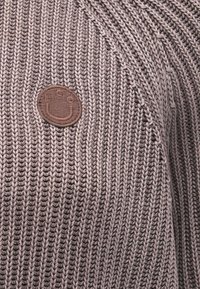 Kootud kampsun summutatud pruunides toonides, millel on tekstureeritud soonikdisain ja ees ümmargune pruunist kangast logoplaat.