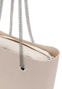 Beżowa torba O z gładką plastikową obudową, srebrnymi łańcuszkowymi uchwytami i kontrastującą beżową, zapinaną na zamek wewnętrzną przegrodą z tkaniny.