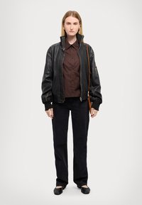 Chaqueta bomber de cuero negro con cuello alto sobre una camisa marrón de botones, combinada con pantalones negros y zapatos planos negros.