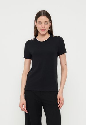 Kvinde med langt brunt hår iført sort kortærmet top med guldknapper på skuldrene og sorte bukser, stående mod hvid baggrund.