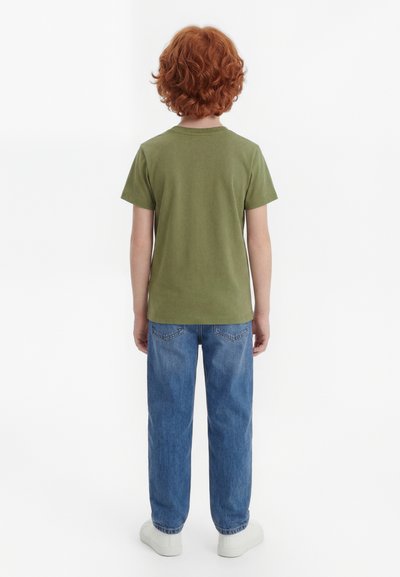 Niño con cabello rizado rojo que lleva una camiseta verde, jeans azules y zapatillas blancas de pie de espaldas sobre un fondo blanco liso.