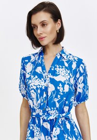 Niebieska sukienka koszulowa w kwiaty z krótkimi rękawami, białym wzorem kwiatowym, kołnierzykiem, guzikami z przodu oraz zawiązywaniem w talii.