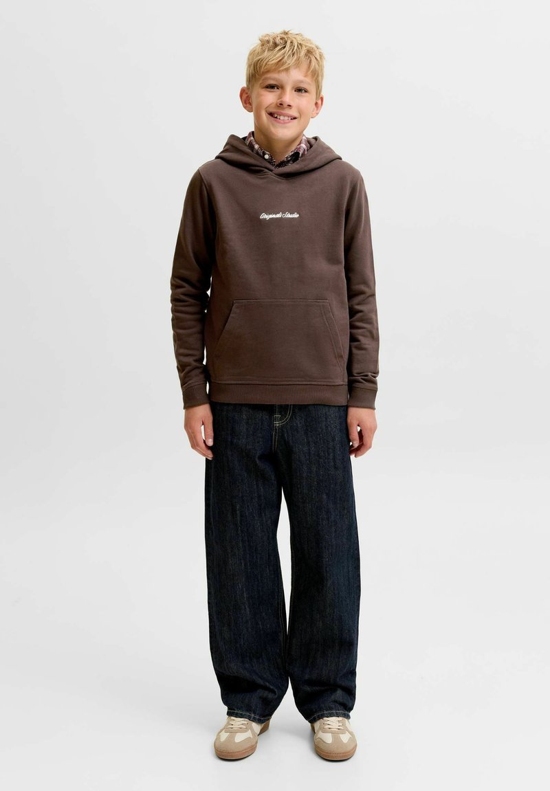 Garçon blond souriant debout, portant un sweat à capuche marron avec poche avant, un jean large foncé et des baskets beiges sur un fond clair uni.