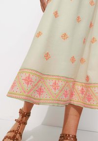 Jupe longueur cheville en tissu beige clair avec des broderies florales roses, présentant une bordure brodée géométrique et un design fluide.