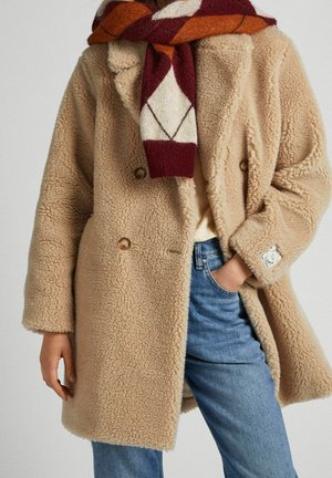 Manteau beige en polaire à double boutonnage porté sur une chemise claire, jean bleu, et écharpe aux motifs rouges, blancs et orange. Main dans la poche.