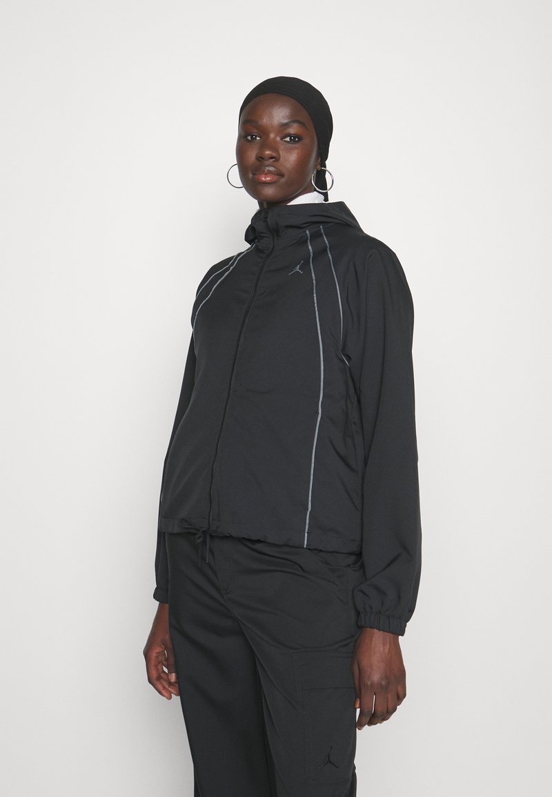 Chaqueta negra con capucha, puños elásticos y dobladillo ajustable; presenta líneas reflectantes en gris y un pequeño logo en el pecho.