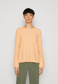 Chemise longue à manches longues orange clair avec un motif floral blanc, col rond et coupe décontractée, associée à un pantalon large vert olive.