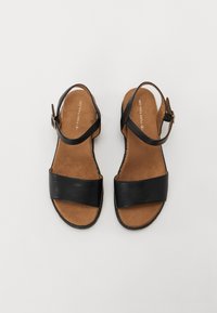 Sandales en cuir noir avec deux larges brides et une fermeture à boucle. Elles présentent une semelle intérieure texturée marron et un design minimaliste.