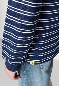 Nieformalny niebieski sweter w paski z białymi poziomymi paskami, teksturowana tkanina, ściągacze u rękawów oraz żółta metka na dole. Noszony z jasnymi szortami dżinsowymi.