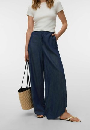 Žena u bijeloj kratkoj majici i širokim tamnoplavim hlačama, drži pletenu torbu boje kože i nosi bež sandale.