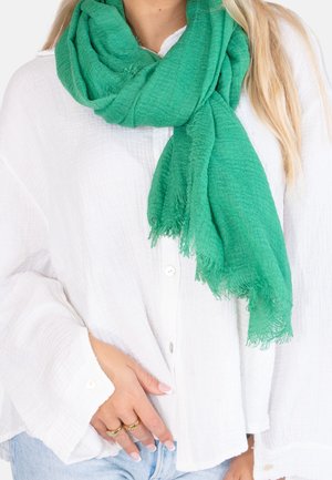 Femme portant une écharpe vert vif sur une chemise blanche fluide boutonnée et un jean bleu clair, avec des bagues en or aux doigts.