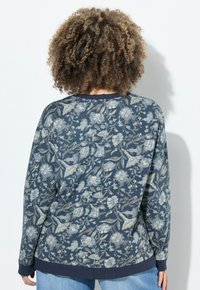 Persona con cabello rizado, vistiendo una blusa de manga larga de patrón floral azul y vaqueros de un azul claro, de frente a una pared blanca.