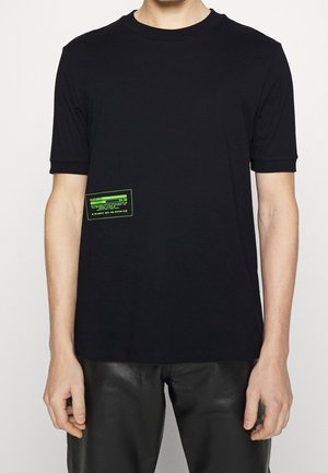Mann trägt ein schlichtes schwarzes T-Shirt mit kurzen Ärmeln und einem kleinen grünen rechteckigen Label auf der unteren linken Seite.