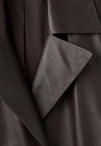 Brązowy skórzany element odzieży o gładkiej teksturze, z nakładającymi się klapami i skośnymi wycięciami. Projekt zawiera subtelne detale przeszycia.