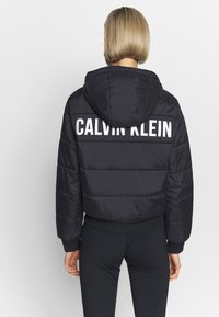 Person med kort, blondt hår iført en sort dunjakke med hætte og "CALVIN KLEIN" trykt i hvidt på ryggen.