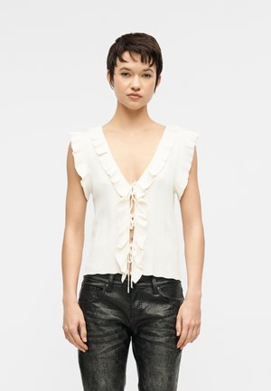 Ung kvinde iført hvid ærmeløs bluse med flæser og V-hals, bundet foran, samt sorte teksturerede læderbukser, stående mod en ensfarvet baggrund.