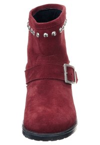 Bottine en daim rouge avec une semelle plate, des clous décoratifs argentés le long du dessus et une bride réglable avec boucle sur le côté.