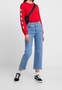 Röd långärmad skjorta med svart logotyp, blå högmidjade vida jeans, svarta sneakers med vita detaljer, och en liten svart crossbody-väska.