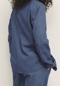 Person med krøllet hår iført en løs blå blazer og matchende bukser, med hånden i lommen, vendt let væk mod en ensfarvet baggrund.