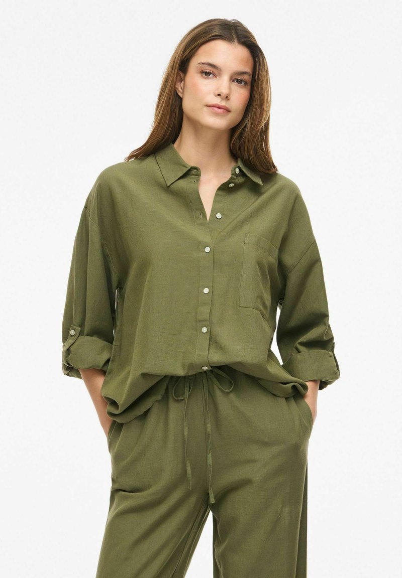 Junge Frau mit langen braunen Haaren, die ein olivgrünes Hemd mit Knöpfen und hochgekrempelten Ärmeln sowie passende Hose mit Kordelzug trägt, Hände in den Taschen.