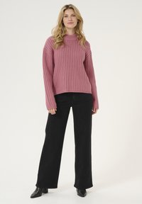 Pink ribbed knit sweater with a high neckline, paired with black wide-leg jeans and ankle boots. Standing model against a white background.