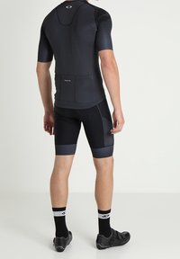 Musta vartalonmyötäinen pyöräilypaita lyhyillä hihoilla, teksturoitu selkäosa ja takatasku. Yhdistetty mustiin pyöräilyshortseihin, polvipituisina. Mustat pyöräilykengät.