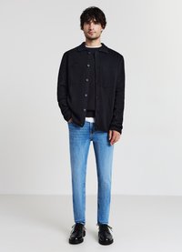 Veste noire à boutons avec deux poches poitrine portée sur un haut sombre, associée à un jean bleu clair et des chaussures noires à lacets. Fond uni.