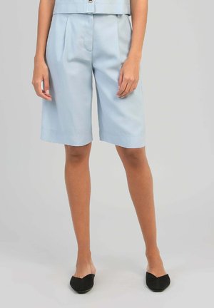 Femme portant un short bermuda bleu clair sur mesure et des mules noires à bout pointu, debout sur un fond neutre.