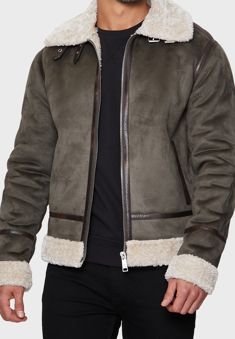 Veste en shearling marron avec un col et des poignets en polaire crème, agrémentée d'accents en cuir et d'une fermeture éclair frontale. Fabriquée en matériau doux et moelleux.
